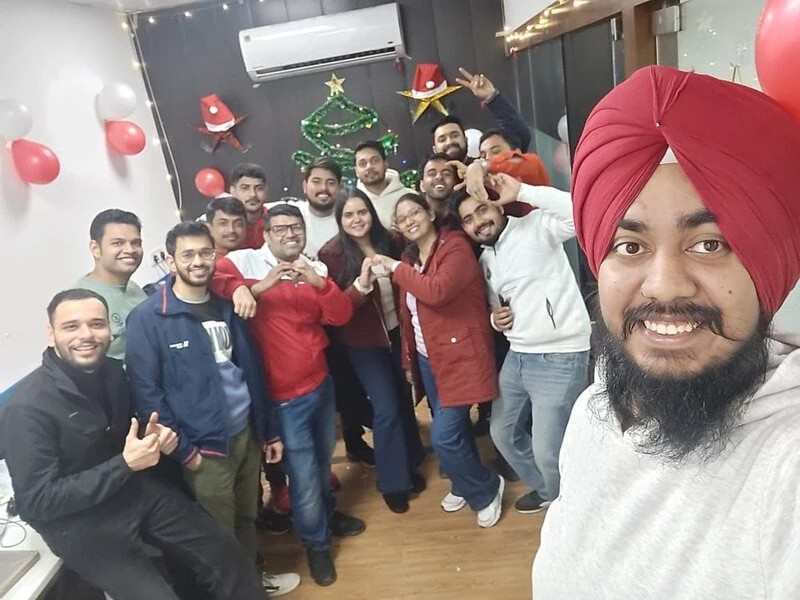 Group selfie of colleagues celebrating Christmas, with red and white balloons and tree decorations in the background.