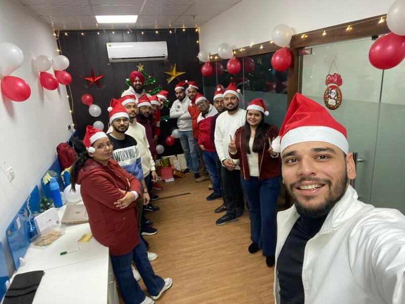 Group photo in a decorated office hallway with red and white balloons, colleagues wearing Santa hats.