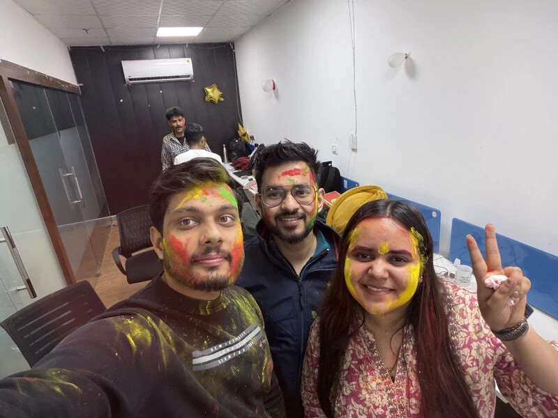 Close-up selfie of three colleagues with red, orange, and yellow Holi powder on their faces, one making a peace sign.
