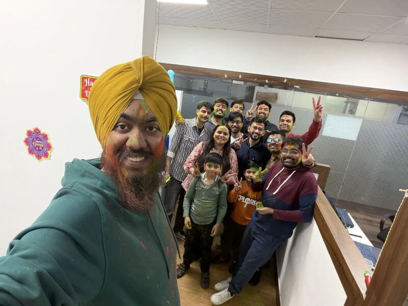 Man in a yellow turban taking a group selfie with colleagues in the office.