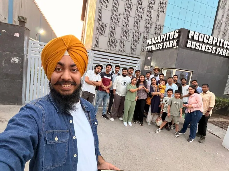 Selfie outside office building: Selfie with a man in an orange turban in the foreground, a large group of colleagues and children behind, outside "Procapitus Business Park."