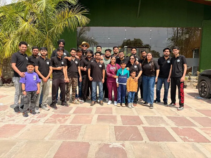 Team with families outdoors: Team members and their families posing together outside a resort building with a green wall and palm trees.