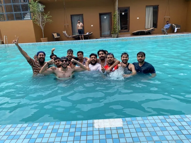 Large pool group cheering: Large group of men swimming and cheering together in a hotel pool, with guest rooms visible behind.