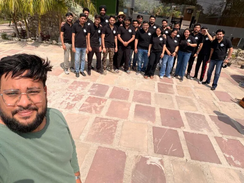 Team selfie with large group: Selfie in front of a group of about 20 colleagues in matching black polo shirts, standing near palm trees outside a building.
